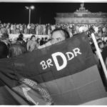 Öffnung der Mauer am 9. November 1989: Ost- und Westbürger Berlins feiern gemeinsam auf der Mauer vor dem Brandenburger Tor den Fall der Mauer. Ein Mann präsentiert die Wiedervereinigung in einer eigens angefertigten Flagge.