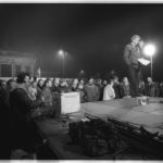 Dezember 1989: ein Moderator umgeben von Schaulustigen wartet auf die "Schalte" in die Abendnachrichten. Im Hintergrund patrouillieren noch DDR-Grenzposten auf der Mauer vor dem Brandenburger-Tor.